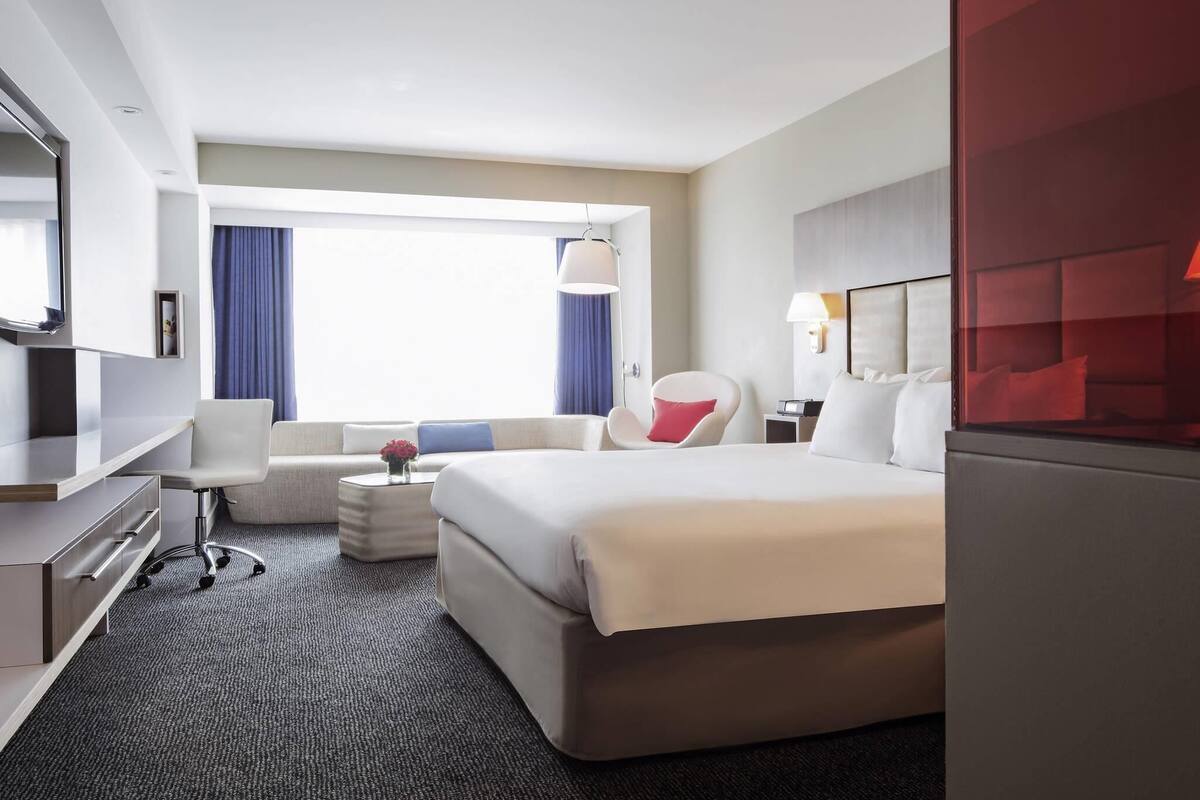 Une chambre d’hôtel avec un lit, un bureau, une chaise, une petite table et une fenêtre avec des rideaux.