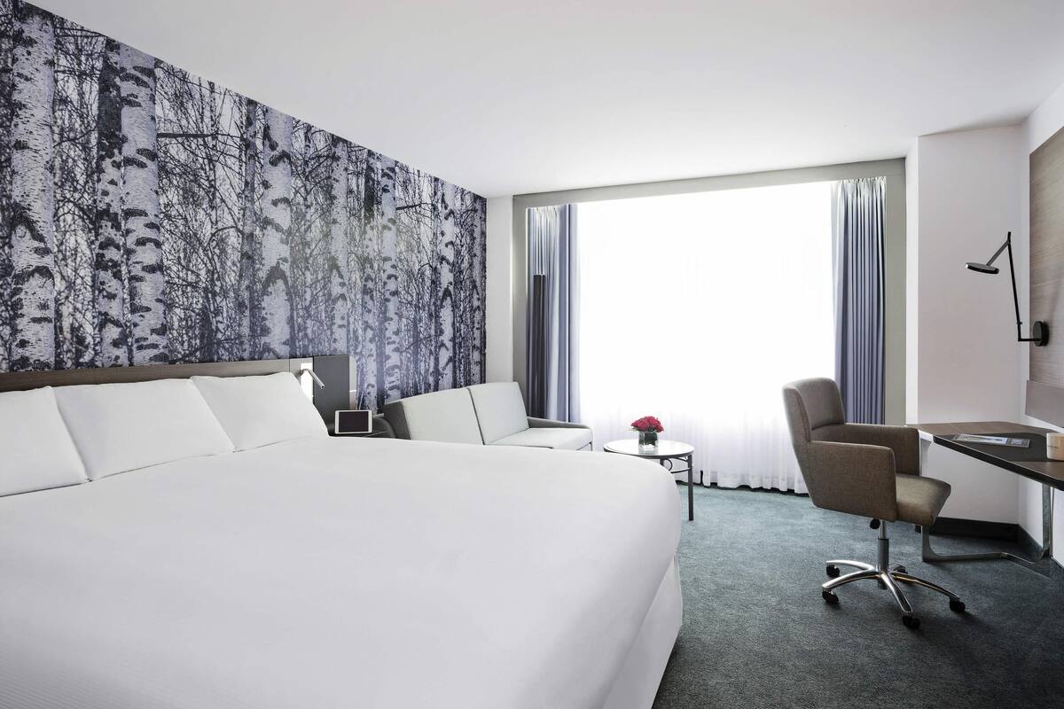 Une chambre d’hôtel avec un grand lit, un bureau, une chaise, une petite table sur laquelle est posé un vase de fleurs, et une grande fenêtre avec des rideaux.