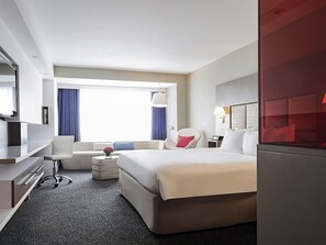 Quarto de hotel com cama, escrivaninha, cadeira, mesinha, luminária e janela com cortinas.