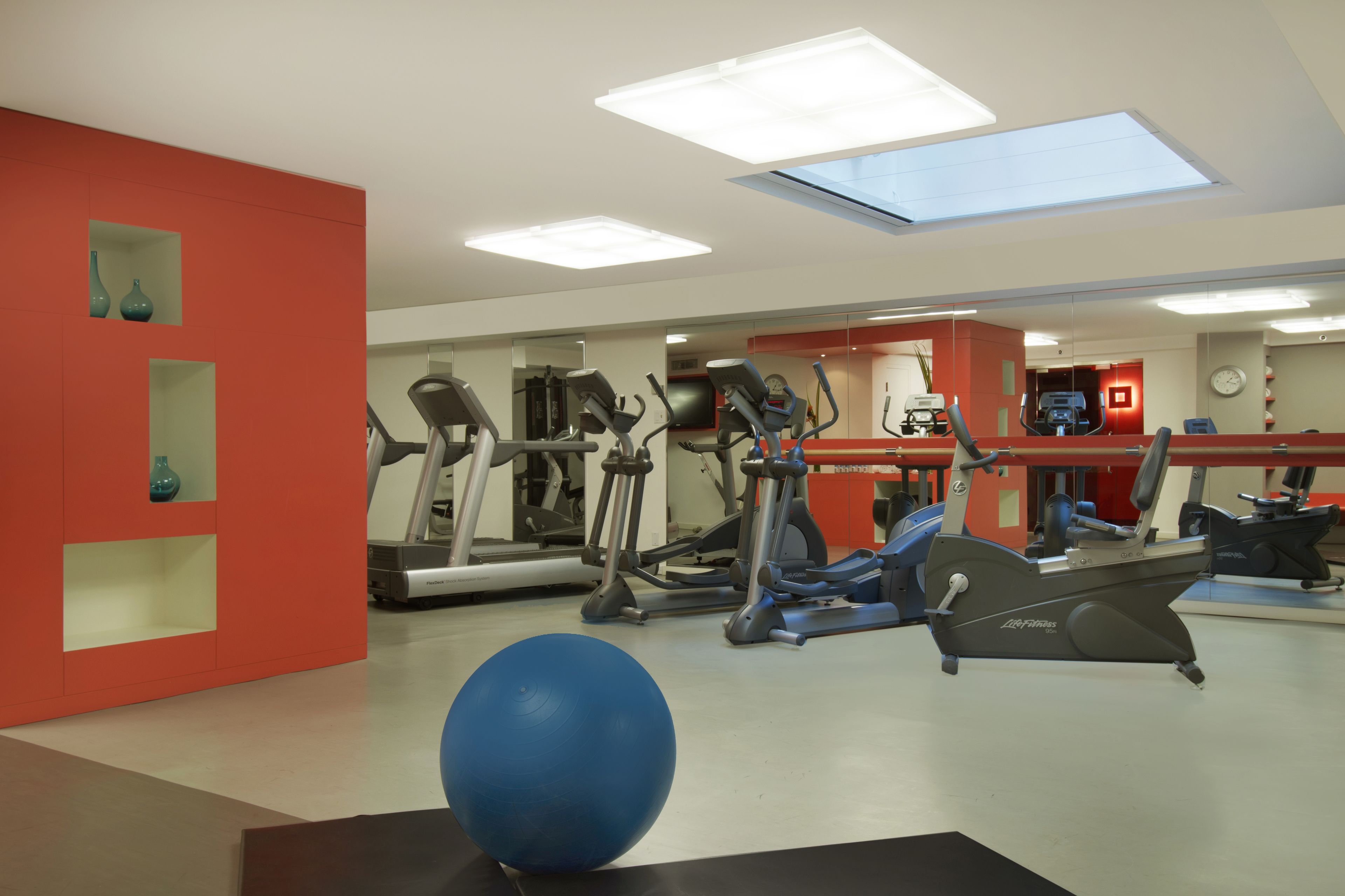 A gym with various exercise machines, a blue exercise ball, and a red wall with shelves.