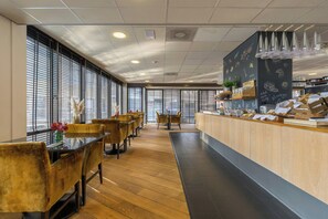 A restaurant with wooden flooring, a counter with food items, and large windows with blinds.