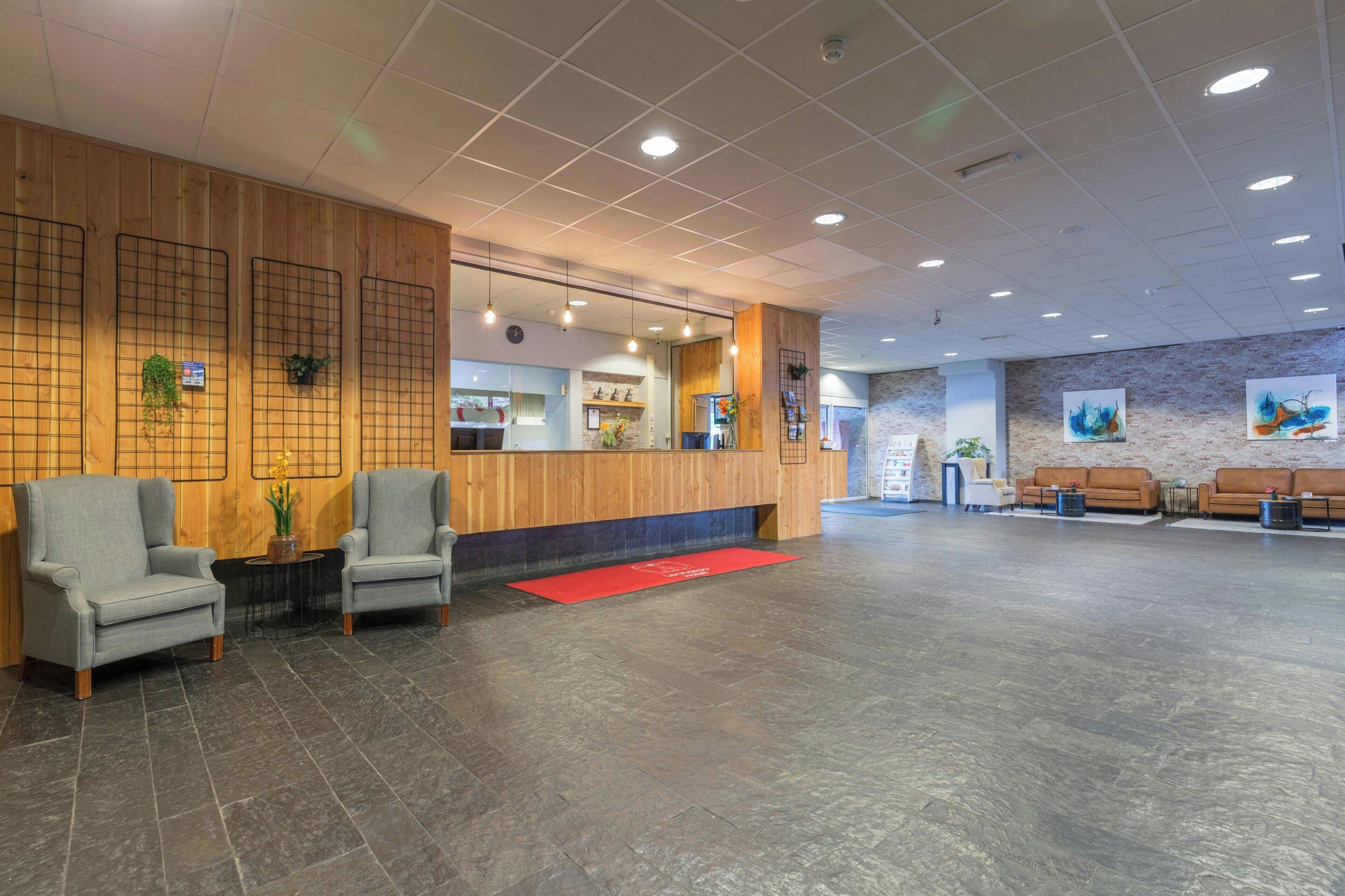 Un hall d’entrée spacieux comprenant un comptoir d’accueil, deux fauteuils, un tapis rouge et un espace détente avec des canapés.