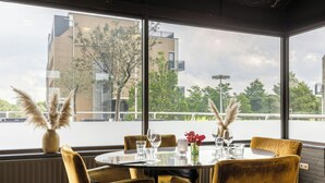 A dining area with a round table, four chairs, and a view of a building and trees through large windows. - Leonardo Hotel Lelystad City Center (Lelystad)