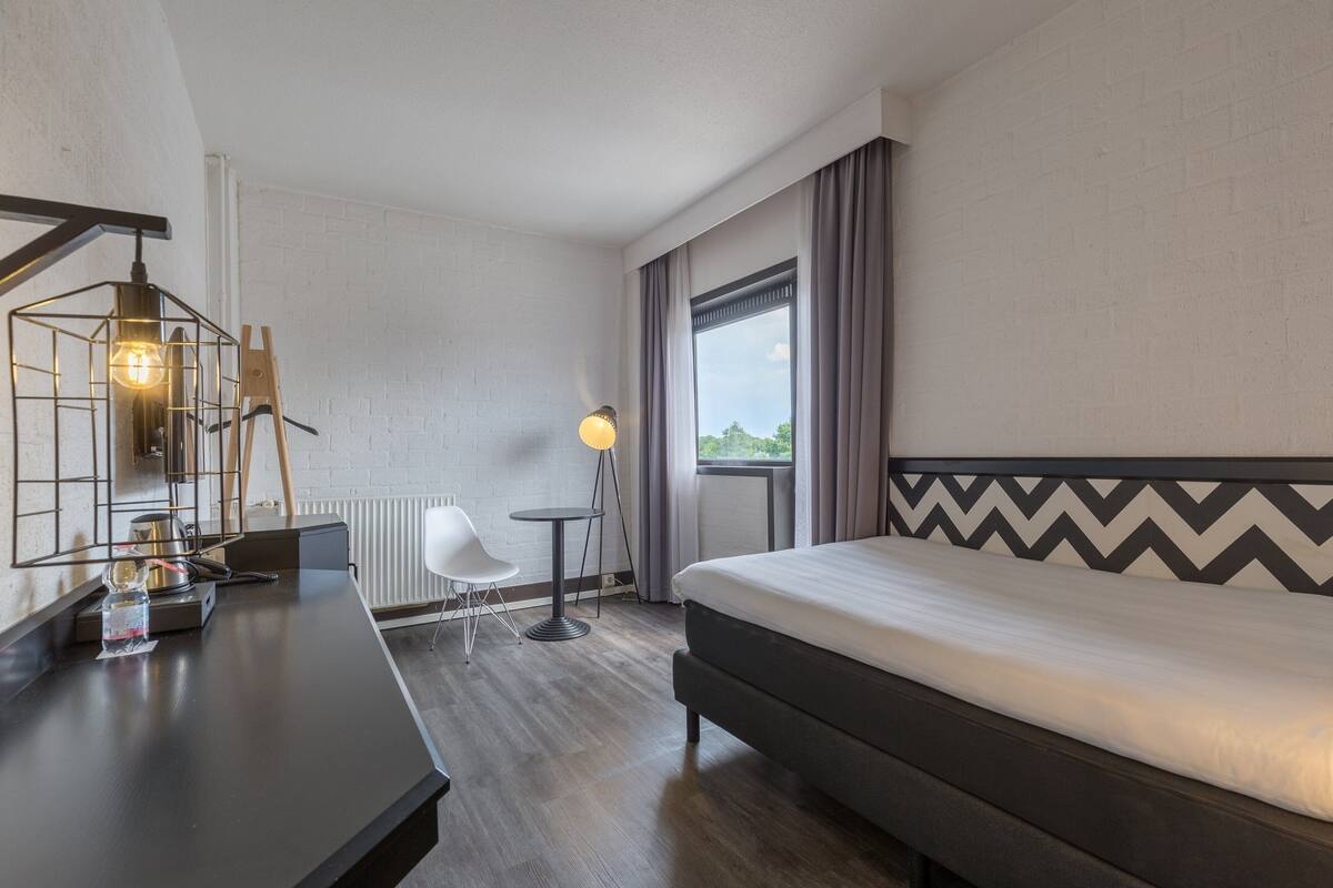 Een hotelkamer met een bed, een bureau, een stoel, een lamp en een raam met gordijnen.