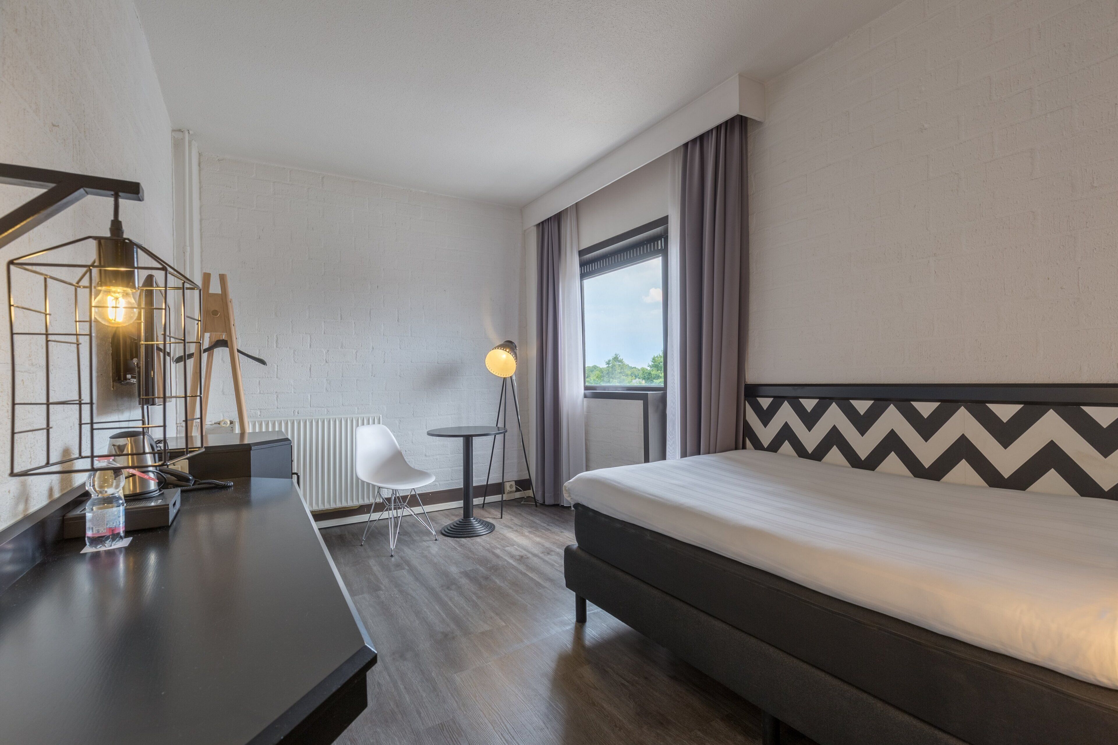 A hotel room with a bed, a desk, a chair, a lamp, and a window with curtains.
