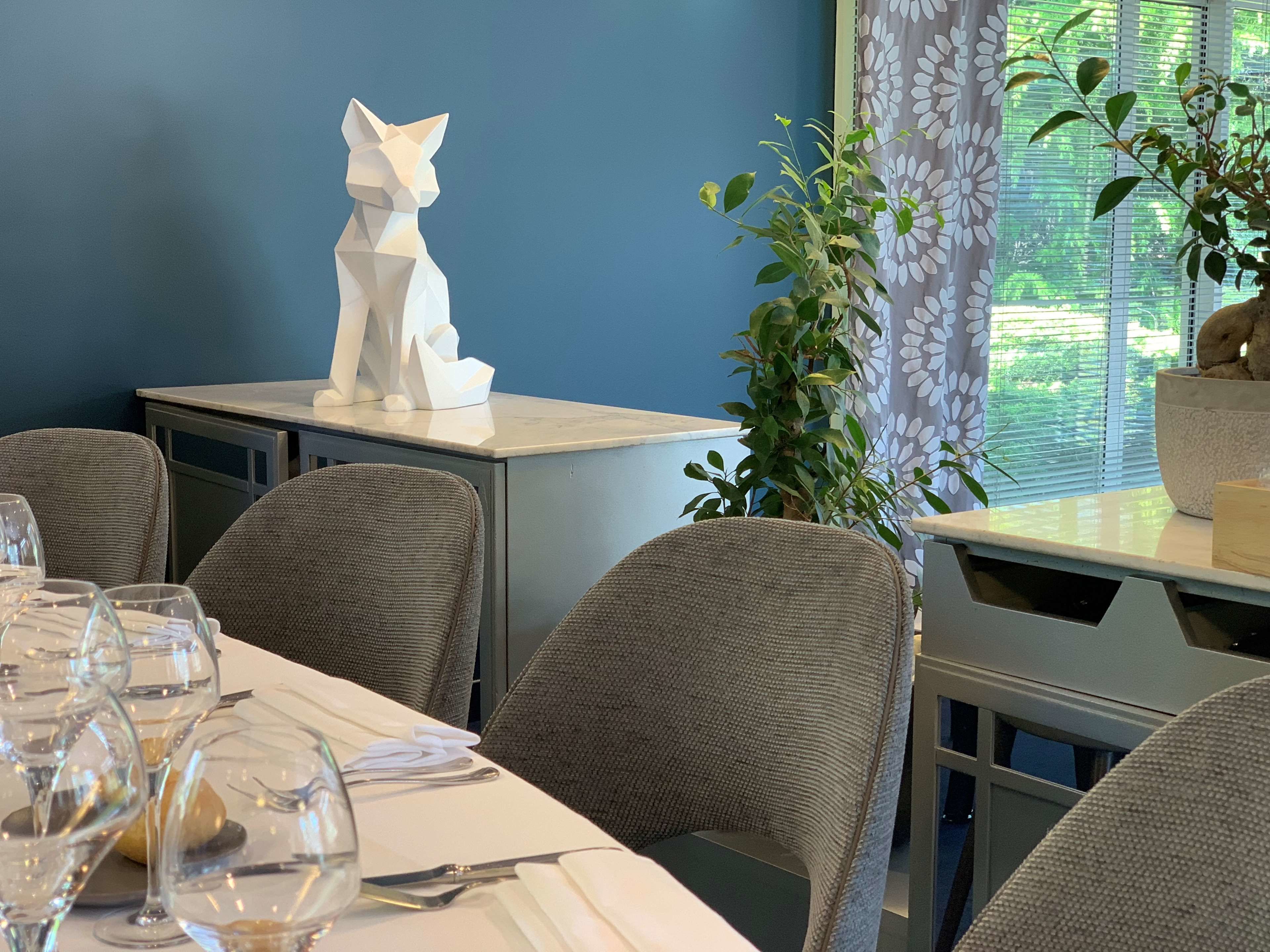 A dining area with a table set for a meal, chairs, a potted plant, and a decorative cat sculpture.
