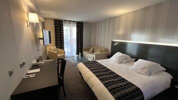 Una habitación de hotel con cama, dos sillones, un escritorio, televisor y ventana con cortinas.
