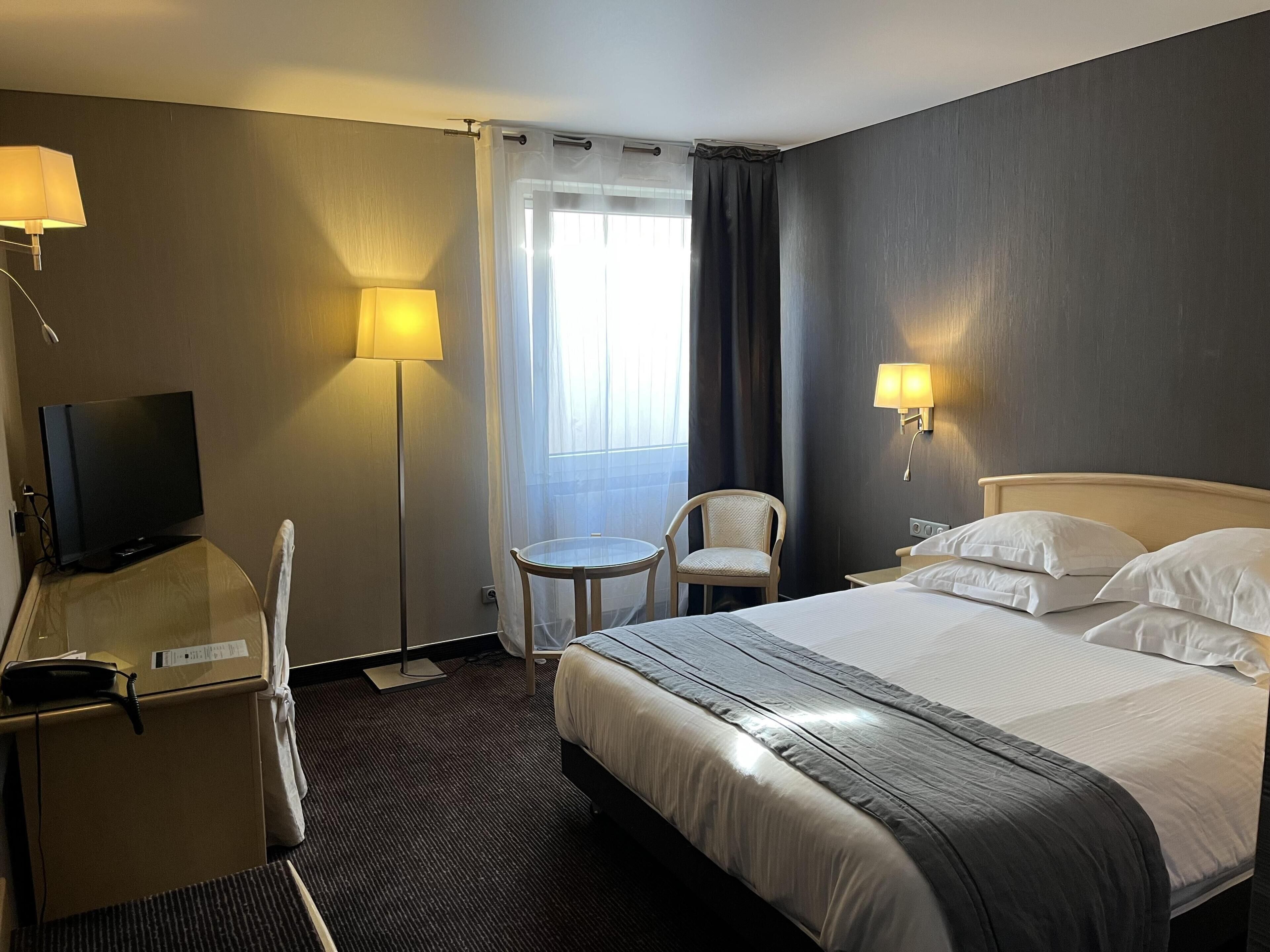 A hotel room with a bed, a desk with a television, a chair, a small table, and a window with curtains.