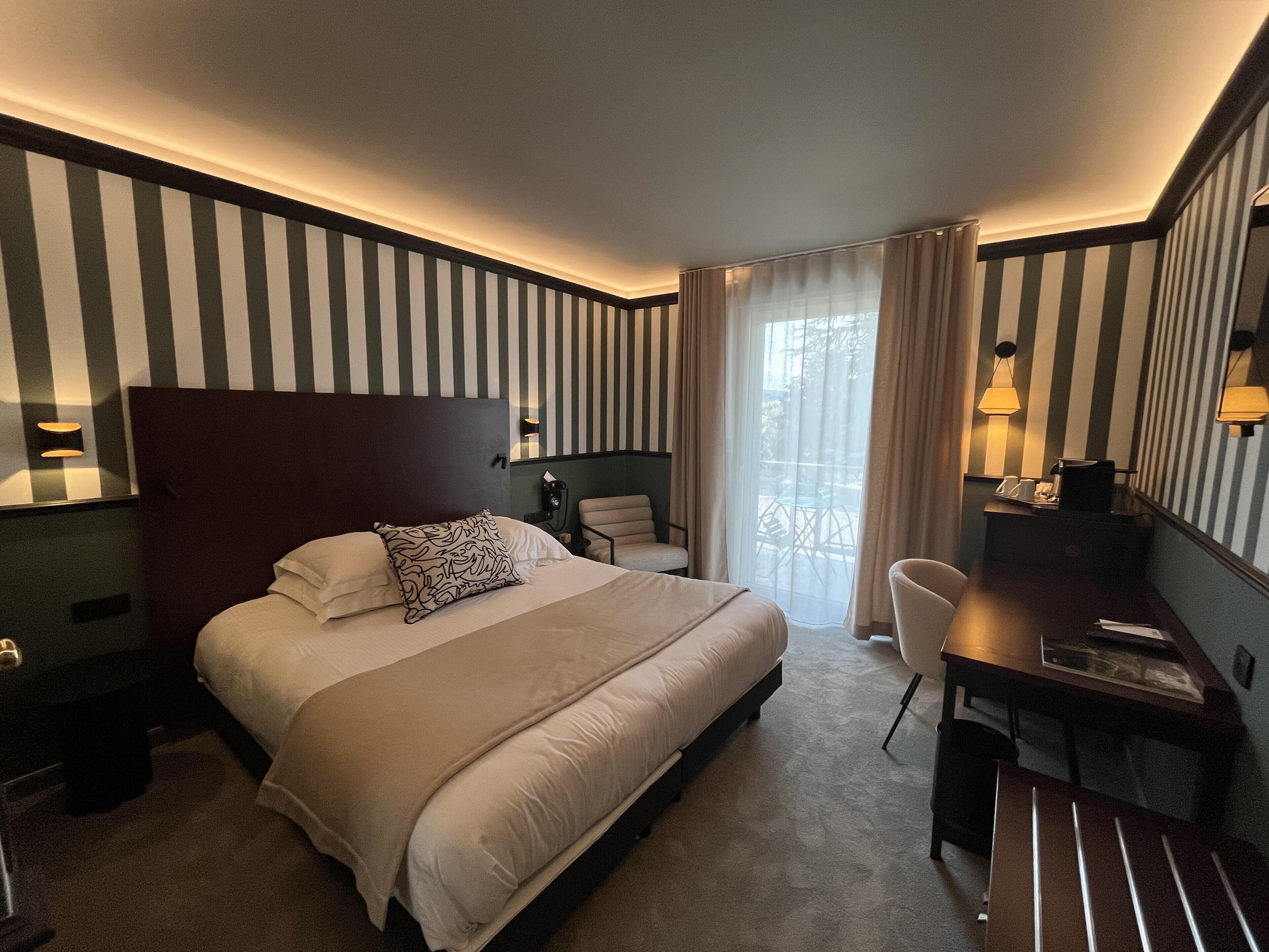 A hotel room with a large bed, a desk, a chair, and a view of the outdoors through a window.