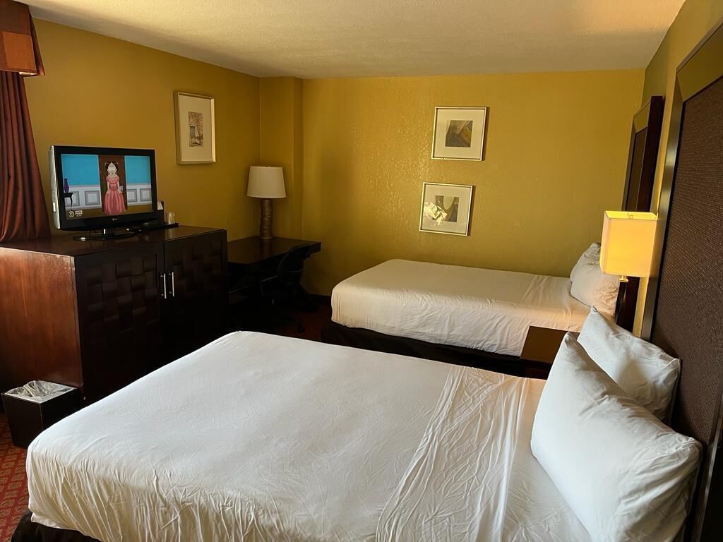 A hotel room with two beds, a desk with a television, and framed pictures on the wall.