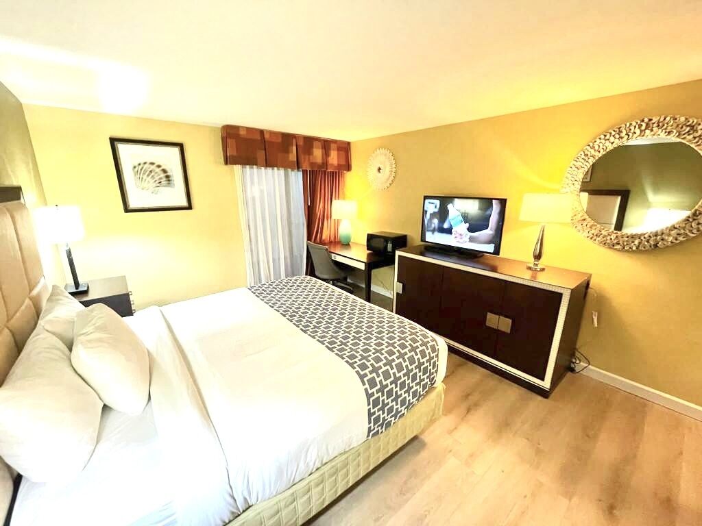 Une chambre d’hôtel avec un lit, un bureau avec une télévision, une commode, un miroir et un tableau encadré au mur.