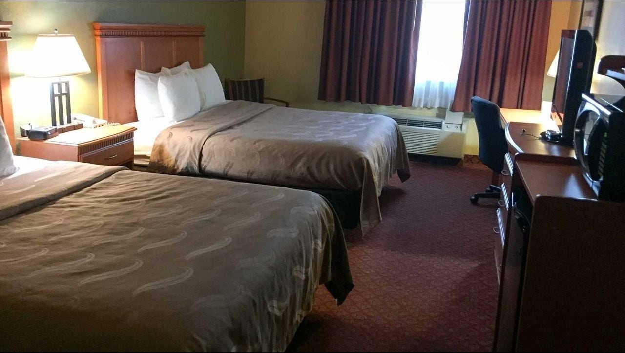 A hotel room with two beds, a desk, a chair, a television, and an air conditioning unit.