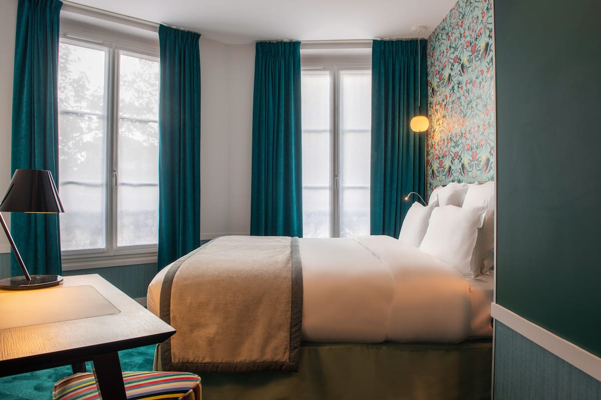 A hotel room with a bed, a desk, a lamp, and a window with curtains.