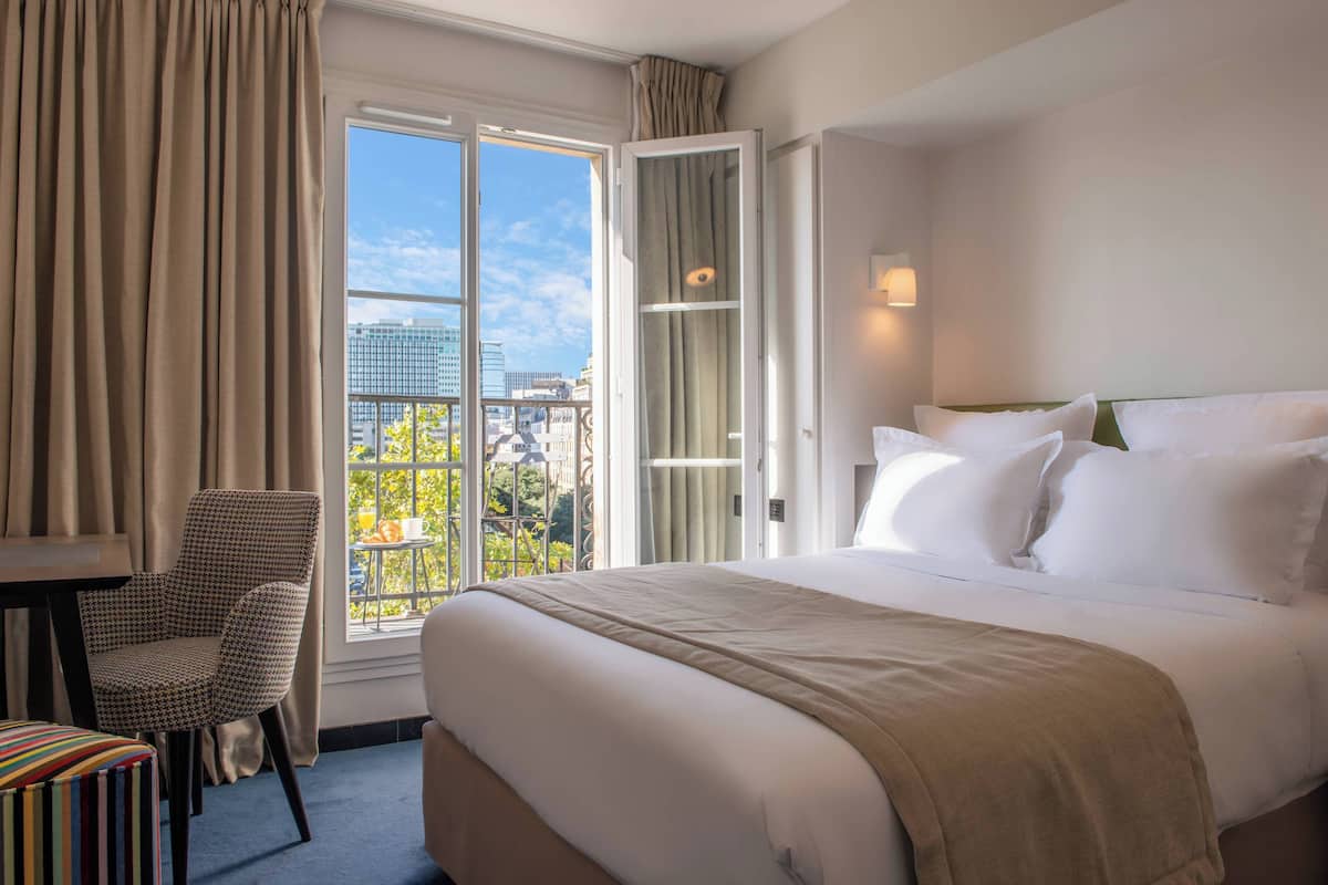 A hotel room with a large bed, a chair, a small table, and a window with a view of the city.