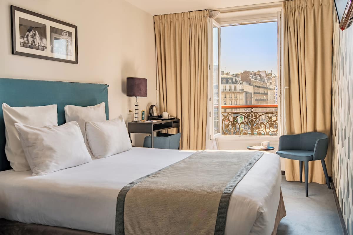 A hotel room with a bed, a desk, a chair, a window with curtains, and a balcony with a railing.