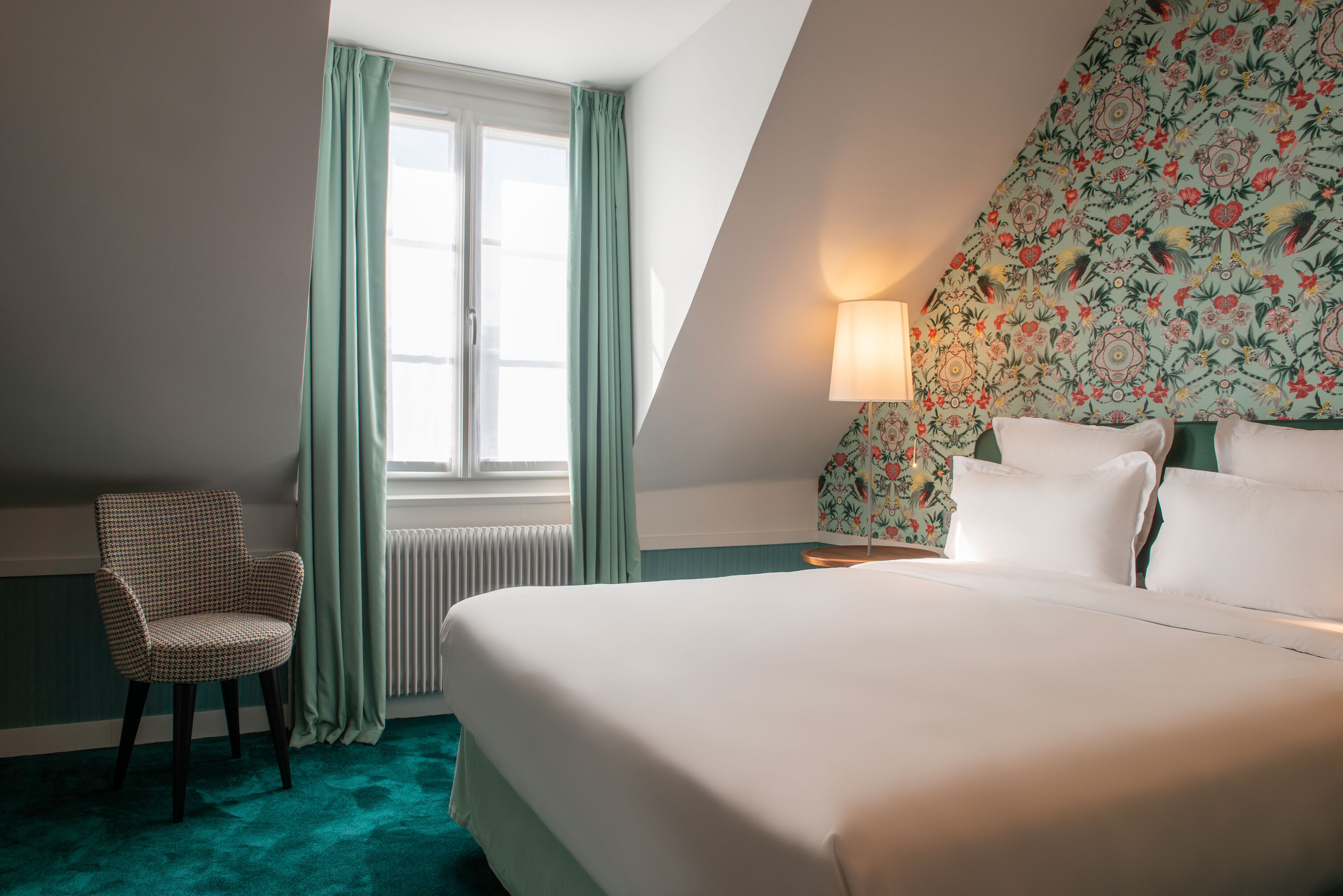 A hotel room with a large bed, a chair, a window with curtains, a radiator, and a floral-patterned wall.
