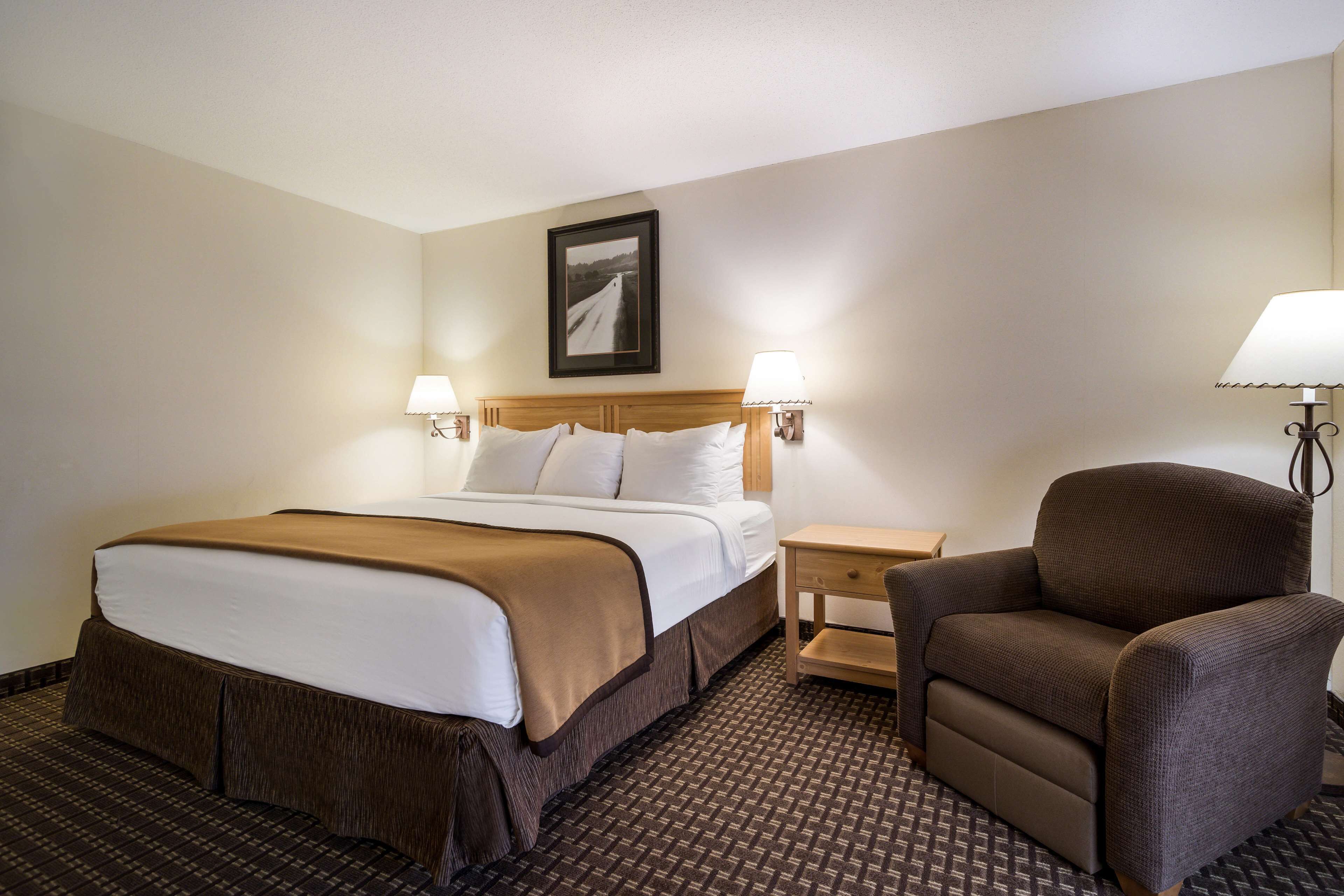 Ein Hotelzimmer mit einem Bett, zwei Nachttischlampen, einem Sessel, einem Nachttisch und einem gerahmten Bild an der Wand.