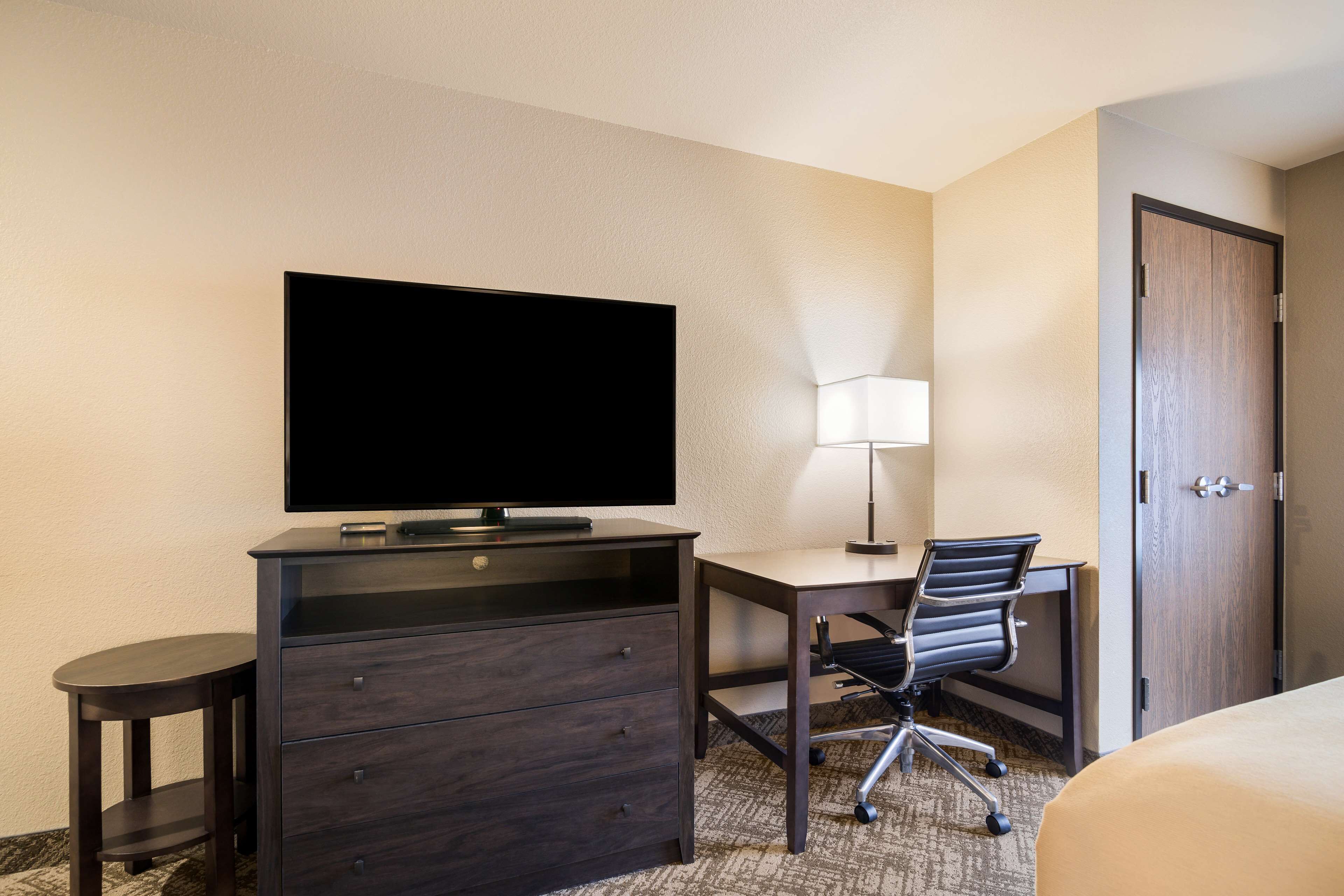 A hotel room with a flat-screen TV, a desk with a chair, a lamp, and a door.