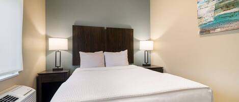 A neatly made bed with white linens, two bedside tables with lamps, a wall-mounted artwork, and a window with blinds.