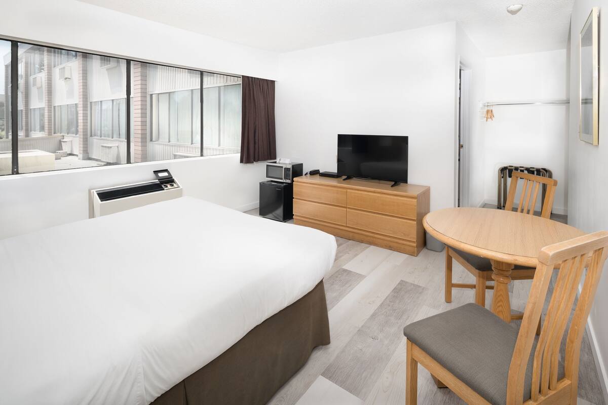 Una habitación de hotel con una cama grande, televisión, cómoda de madera, mesa de comedor con sillas y ventana con cortinas.
