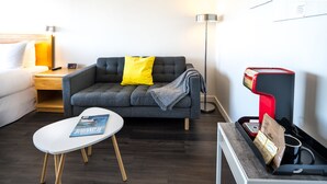 A modern hotel room with a grey sofa, a white table, a floor lamp, and a coffee machine. - Cassandra Hotel (Vancouver)