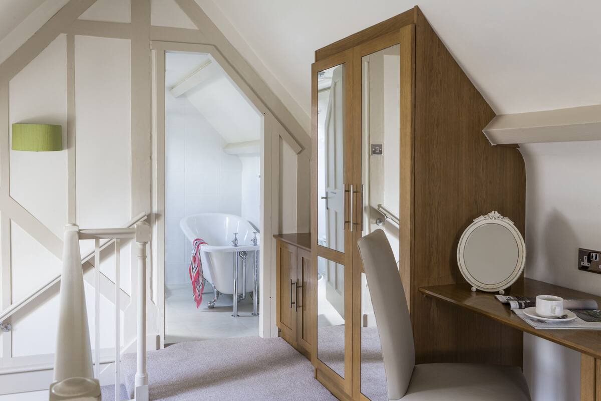 A bedroom with a wooden wardrobe, a mirror, a bedside table, and a view of a bathroom with a bathtub.
