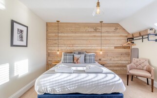 Un dormitorio con pared de listones de madera, cama con ropa blanca, una silla y una lámpara colgante.