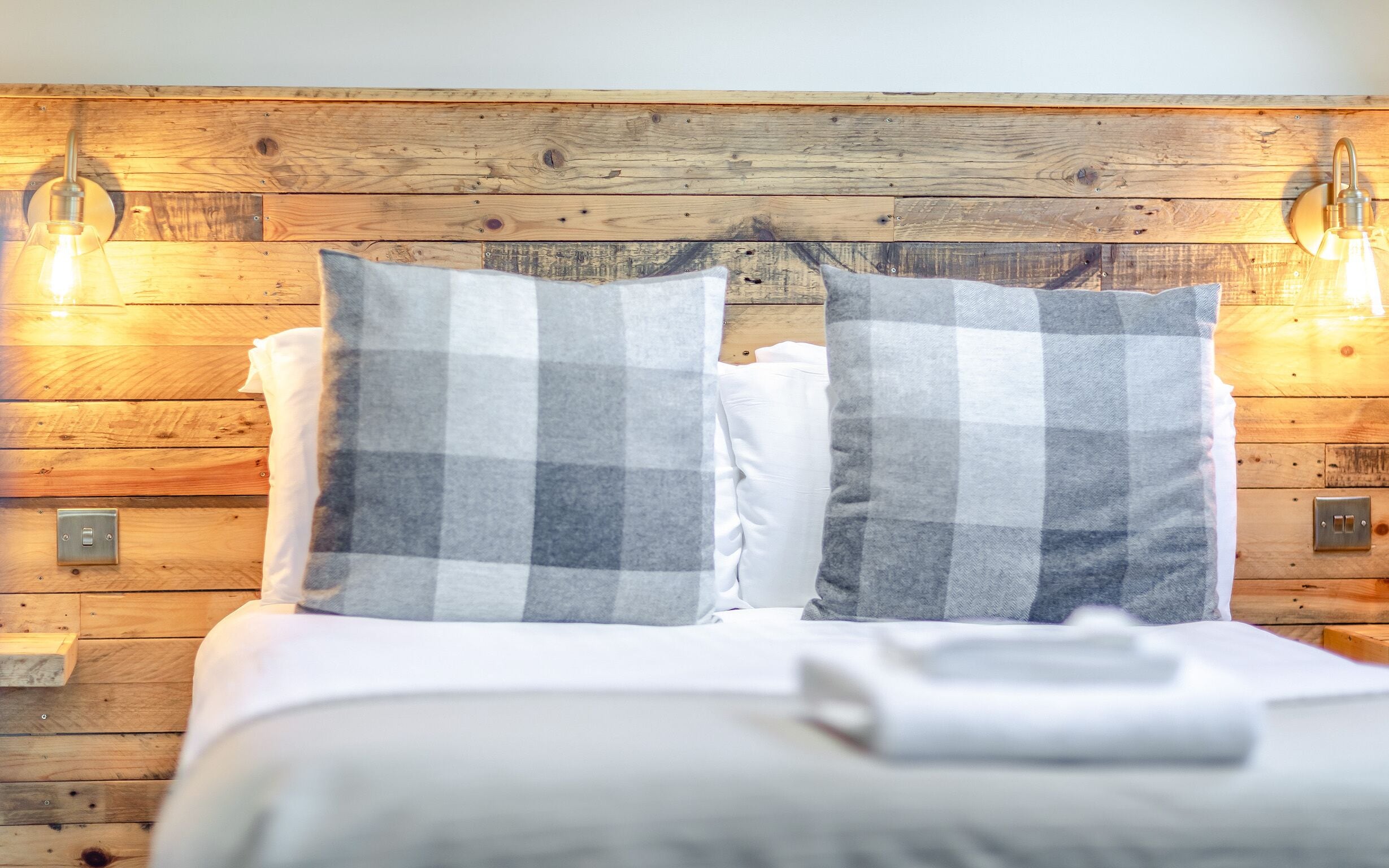 A bed with a checkered pillow and a white pillow, set against a wooden wall with two wall-mounted lights.