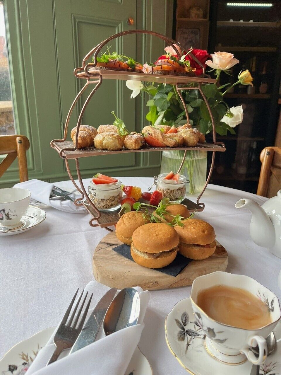 Un soporte escalonado para postres con pasteles y sándwiches, una taza de café y un jarrón con flores sobre una mesa.
