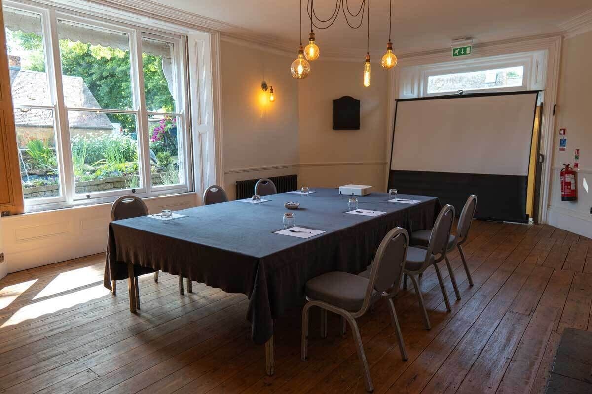 A conference room with a long table, chairs, a projector screen, and a window with a view of greenery.