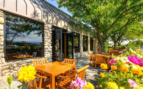 A restaurant with stone walls and large windows, outdoor wooden tables and chairs, and a garden with colorful flowers.