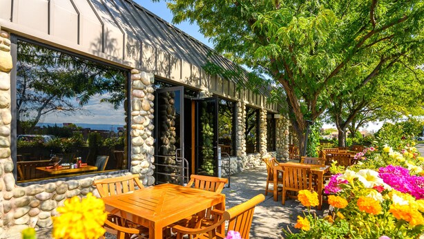 A restaurant with stone walls and large windows, outdoor wooden tables and chairs, and a garden with colorful flowers. - Grand Vista Hotel (Grand Junction)