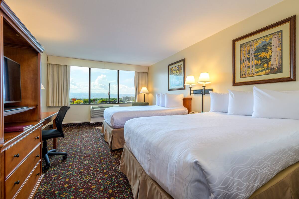 A hotel room with two beds, a desk with a chair, a TV, and a large window with a view.