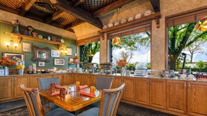 A dining area with a wooden table and chairs, a kitchen counter with various items, and a view of trees through large windows. - Grand Vista Hotel (Grand Junction)