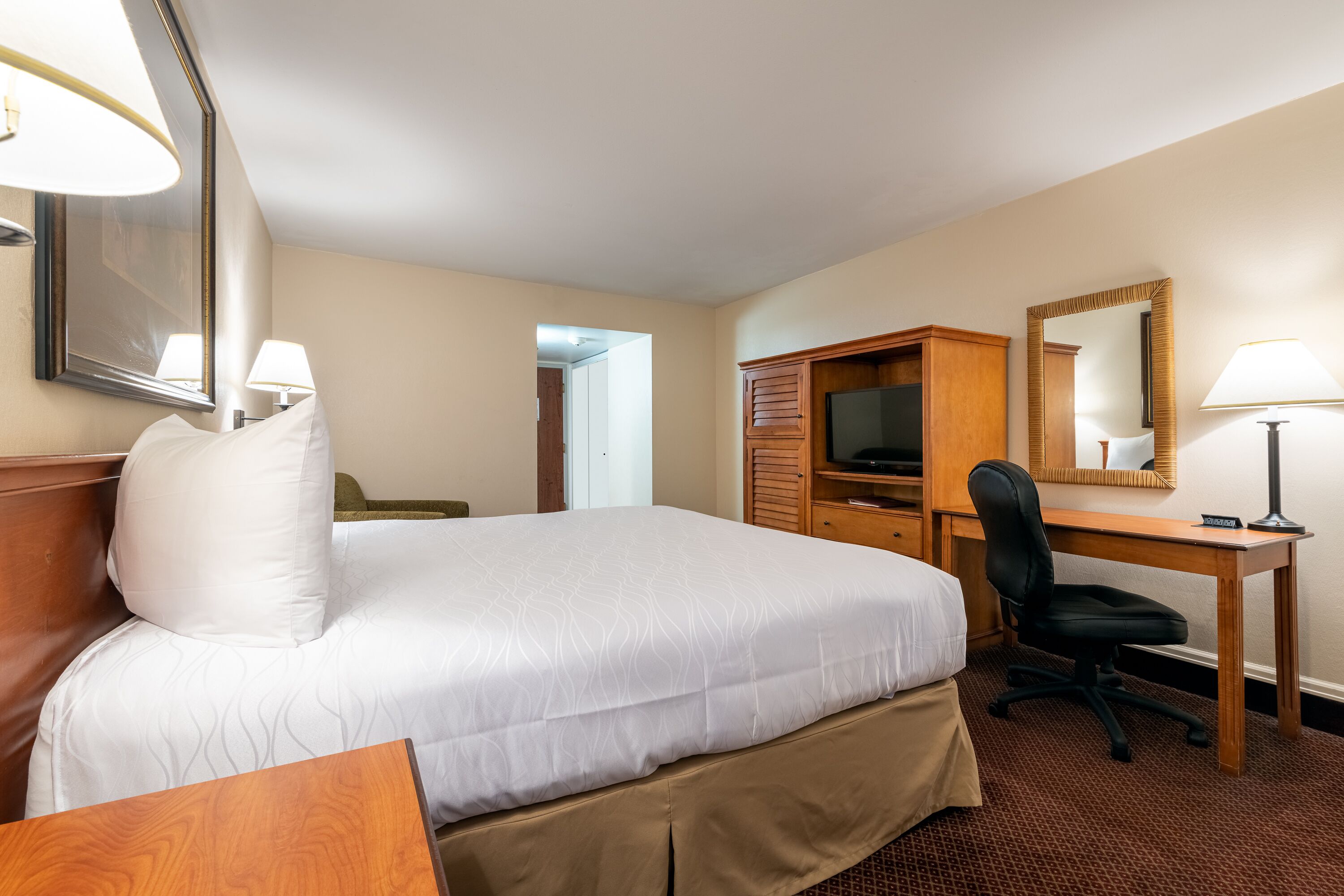 A hotel room with a large bed, a wooden desk, a black office chair, a television, and a mirror.