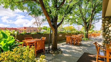 An outdoor patio area with wooden tables and chairs, surrounded by greenery and a stone wall.