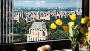 Ein Essbereich mit einem für zwei gedecktem Tisch, einer Vase mit gelben Blumen und Blick auf eine Stadtlandschaft, wobei ein markantes Gebäude im Vordergrund steht.