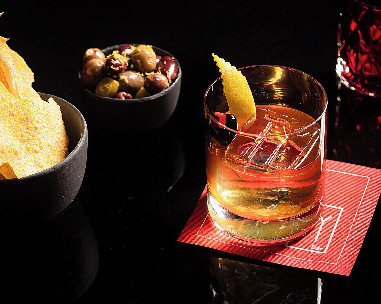 A glass of amber-colored beverage with a lemon peel garnish, accompanied by a bowl of olives and a piece of bread on a dark surface.