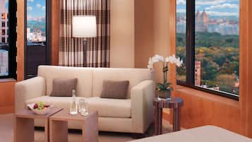 A modern hotel room with a sofa, a small table with a fruit bowl and glasses, a lamp, and a view of a cityscape.