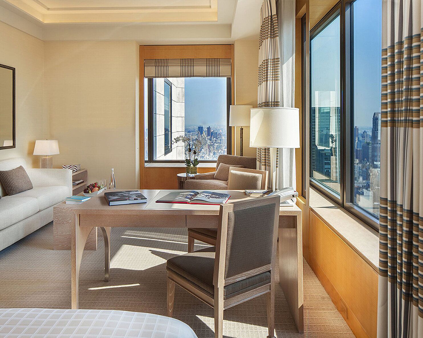 A hotel room with a large window offering a city view, a dining table with chairs, a sofa, and a side table with a lamp and decorative items.
