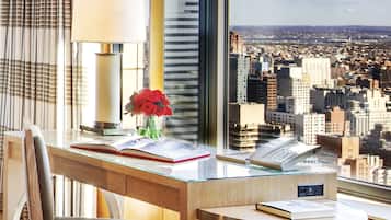 A modern hotel room with a desk, a lamp, a vase with flowers, and a view of a cityscape through large windows.