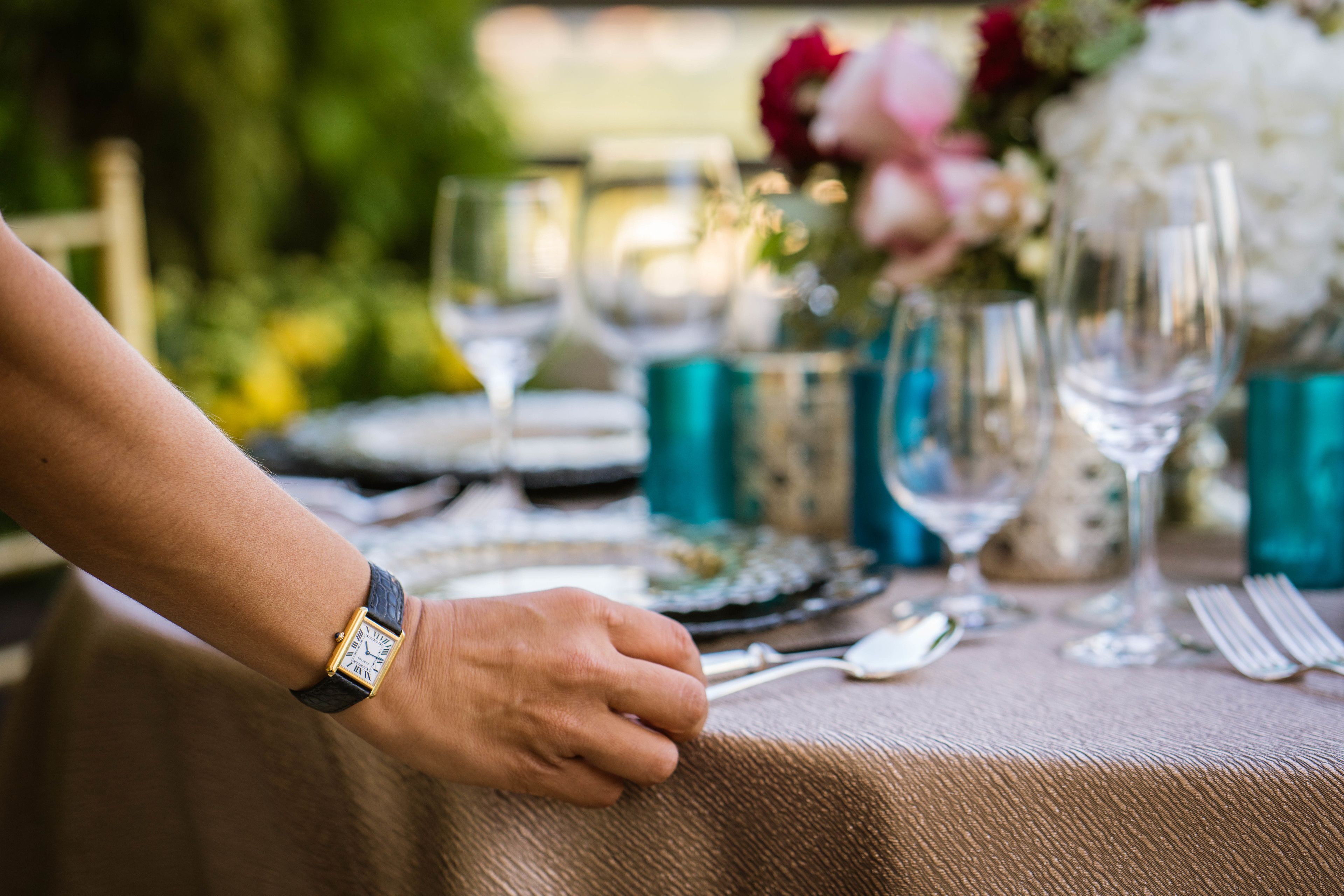 Una mesa dispuesta con un reloj de oro en la muñeca, un tenedor, una cuchara y una copa.