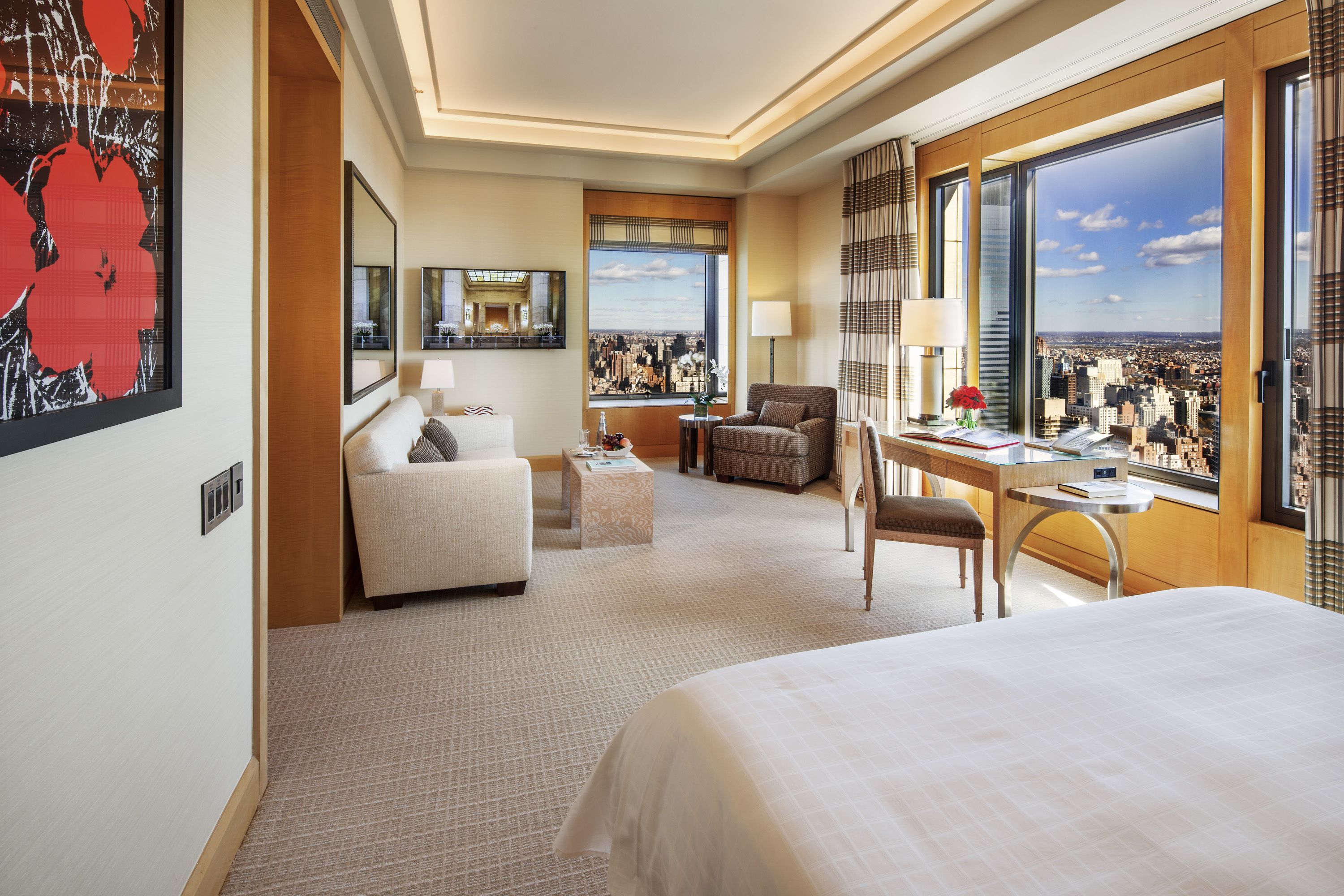 Habitación de hotel con una cama grande, un área de estar con sofá y sillón, un escritorio con silla, un ventanal con vistas a la ciudad y un cuadro en la pared.