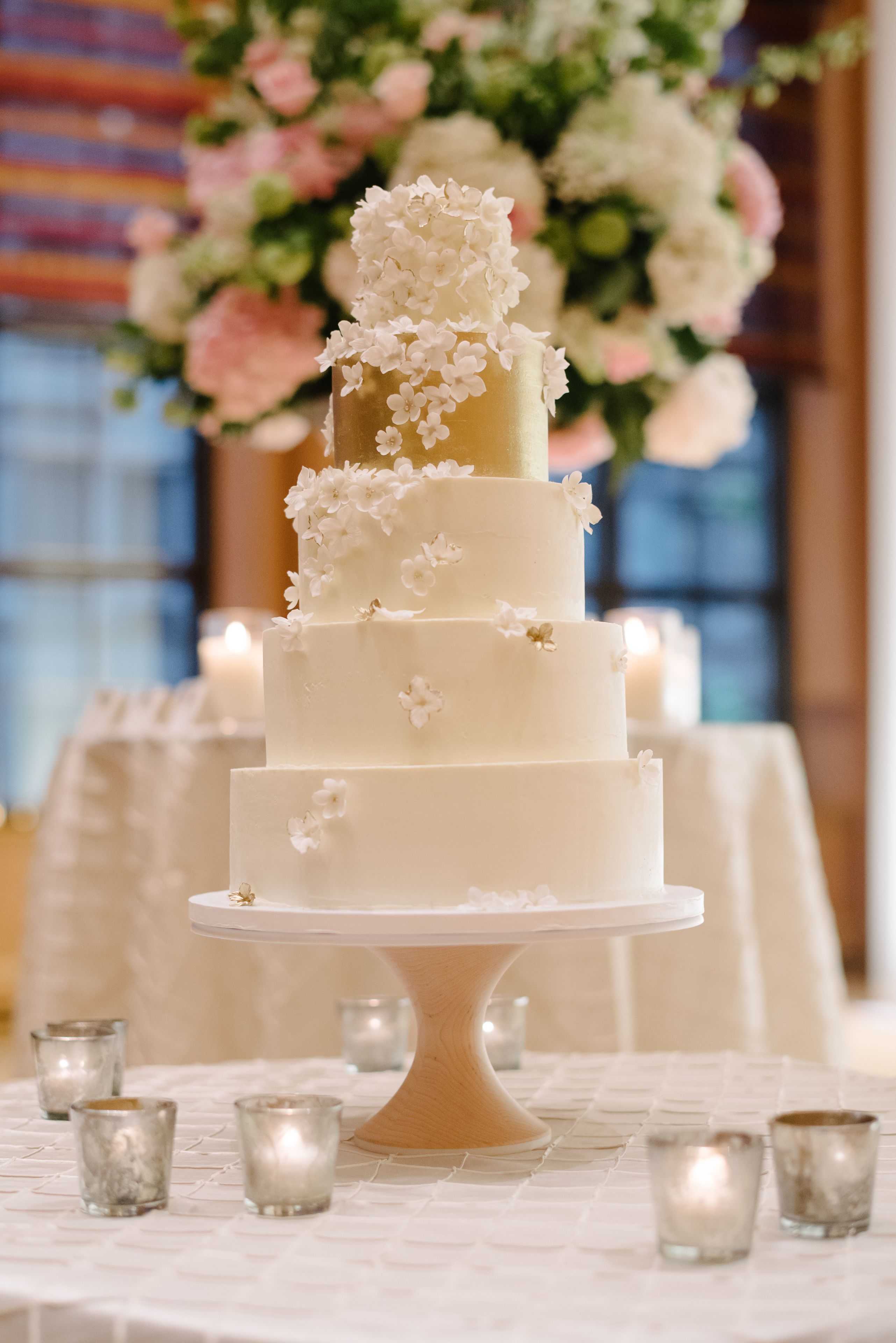 Un gâteau de mariage à plusieurs étages, posé sur un piédestal blanc, entouré de bougies et de petits photophores en verre, le tout sur une nappe.