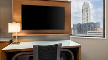 A hotel room with a large flat-screen TV mounted on a wooden panel, a desk with a chair, and a window offering a cityscape view.