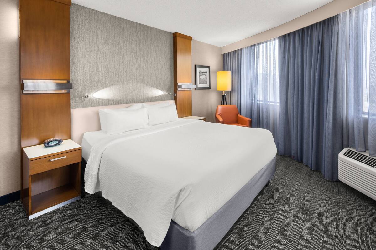 Een hotelkamer met een groot bed, een nachtkastje met een lamp, een stoel en een raam met gordijnen.