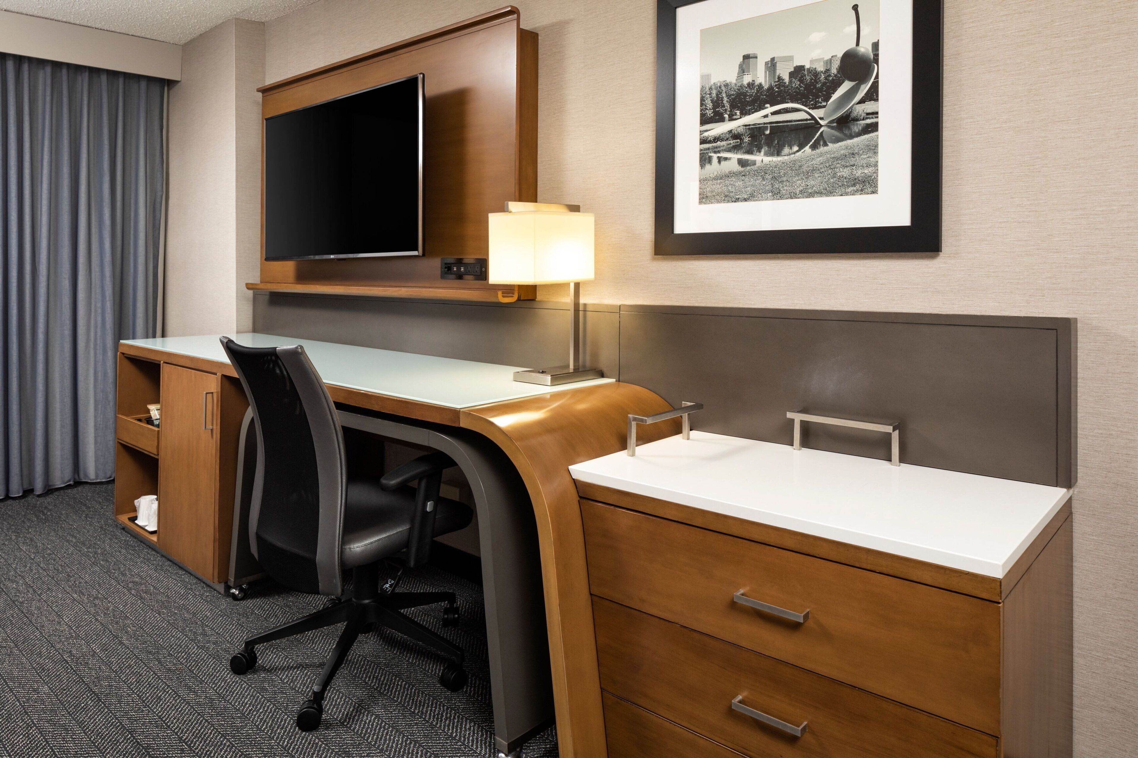 A hotel room with a desk, chair, TV, lamp, and a framed picture on the wall.