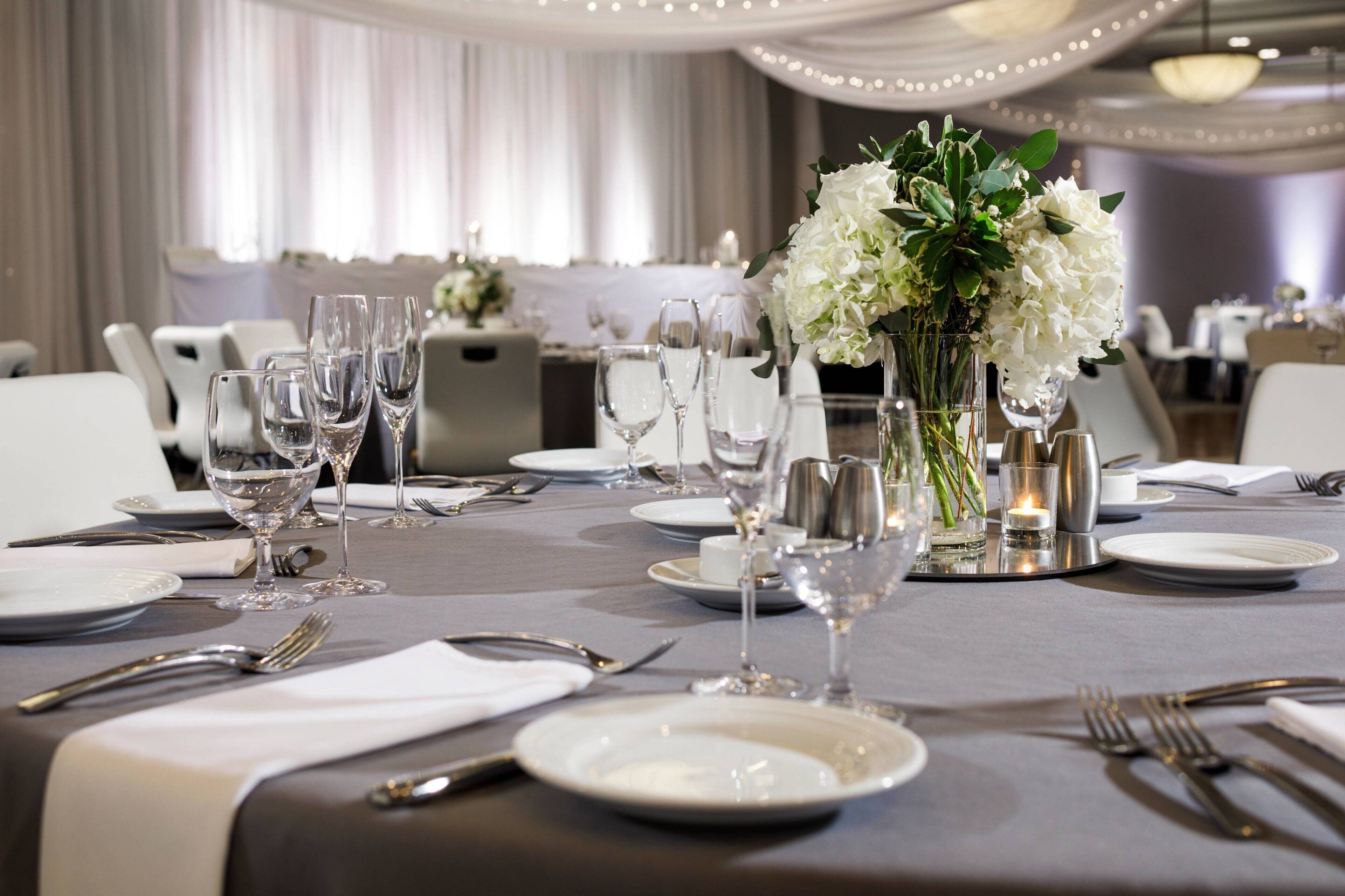 A dining table set with white plates, silverware, and clear glasses, with a centerpiece of white flowers and candles.