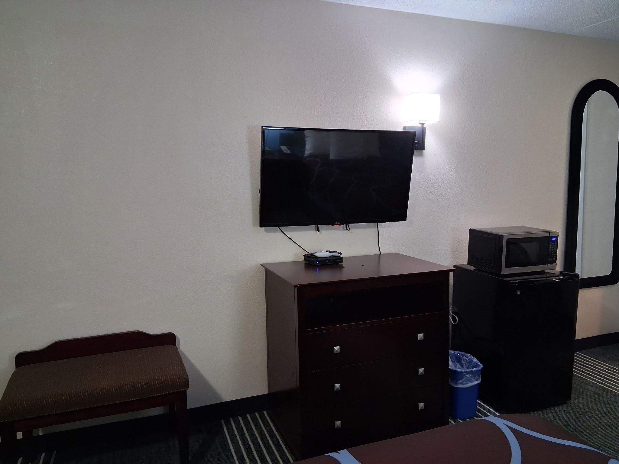 A hotel room with a flat-screen TV mounted on the wall, a small desk with a microwave, a trash bin, and a bench.