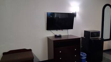 A hotel room with a flat-screen TV mounted on the wall, a small desk with a microwave, a trash bin, and a bench.